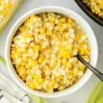 A bowl of fried corn on the table with ears of corn around it and a spoon in the bowl.