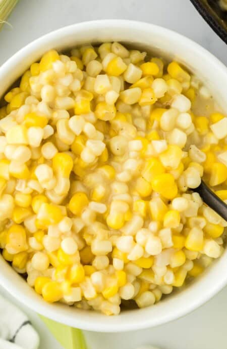 A bowl of fried corn on the table with ears of corn around it and a spoon in the bowl.
