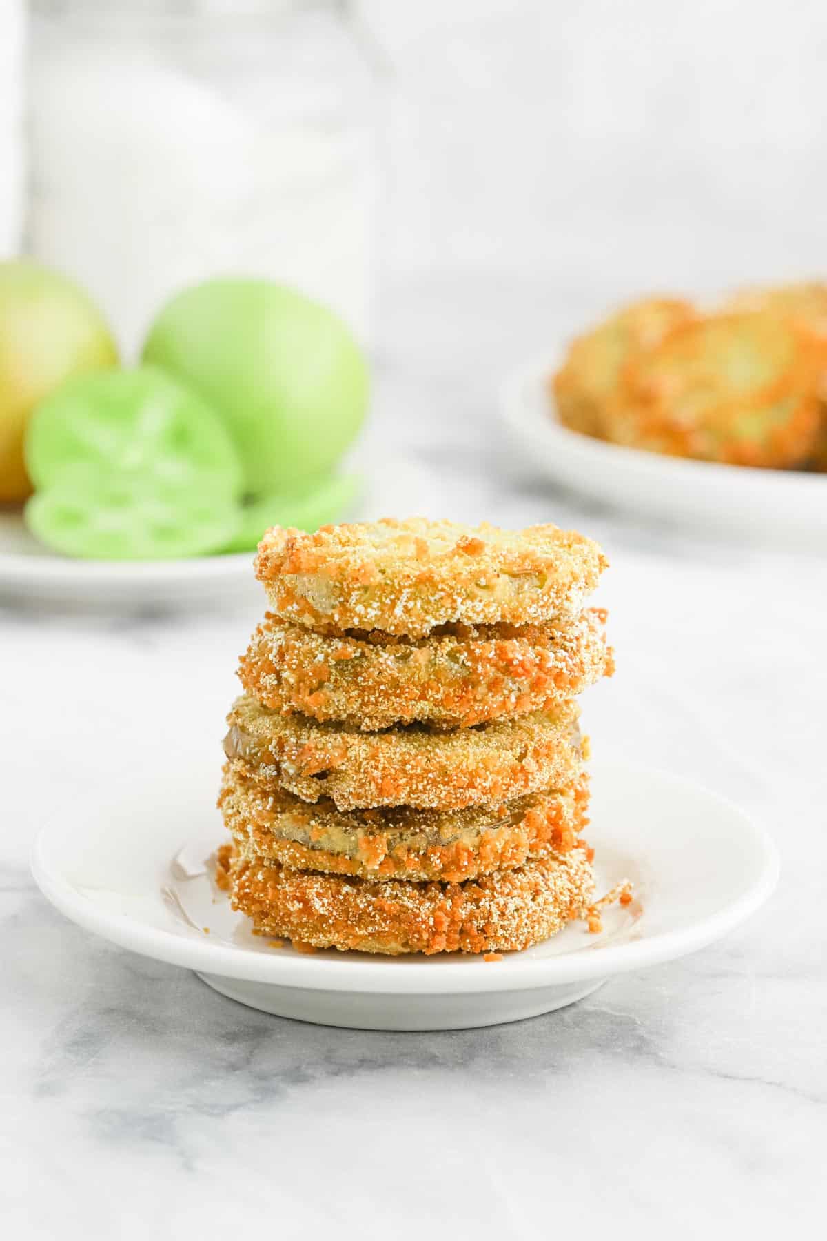 A stack of Southern fried tomatoes on a small white plate with a platter in the background of green tomatoes and more fried.