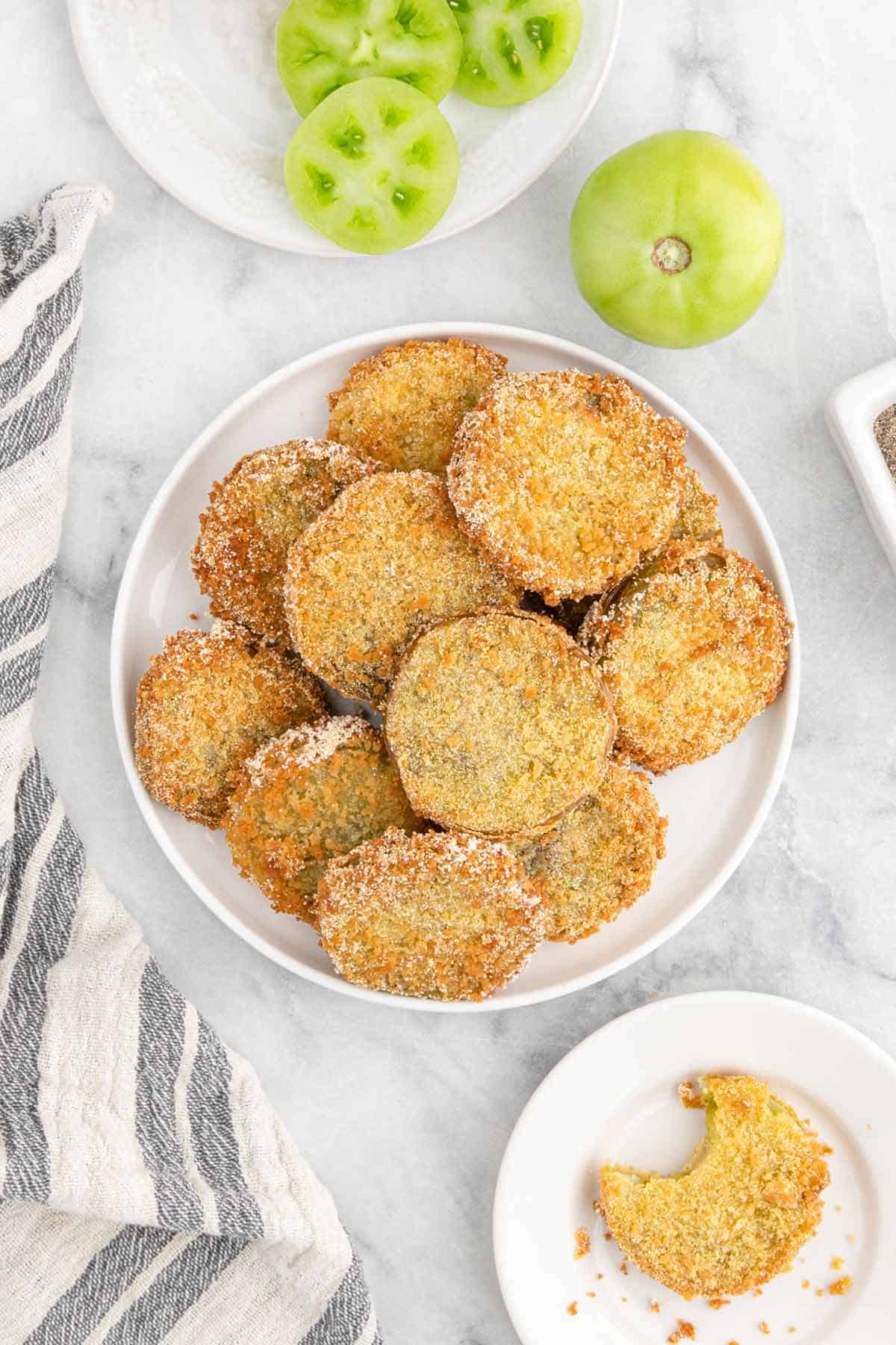 crispy fried green tomatoes stacked up on a plate with a small plate to the side with a fried tomato with a bite missing.