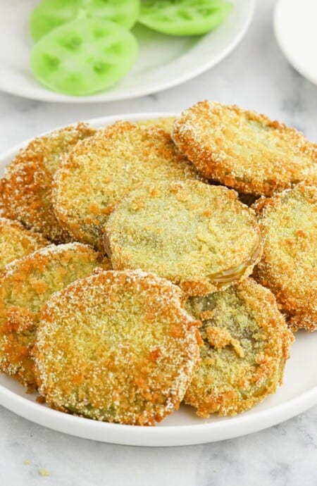 A plate of fried green tomatoes on the table with raw green tomatoes on a plate in the background.