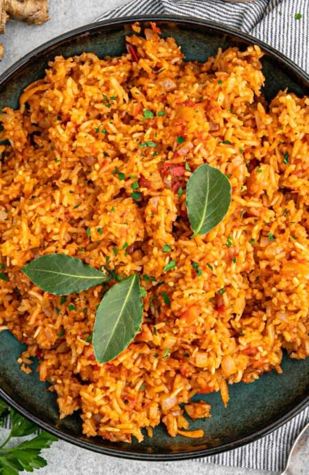 A dark colored bowl of jollof rice recipe on the table garnished with fresh bay leaves and ingredients on the table around the bowl.