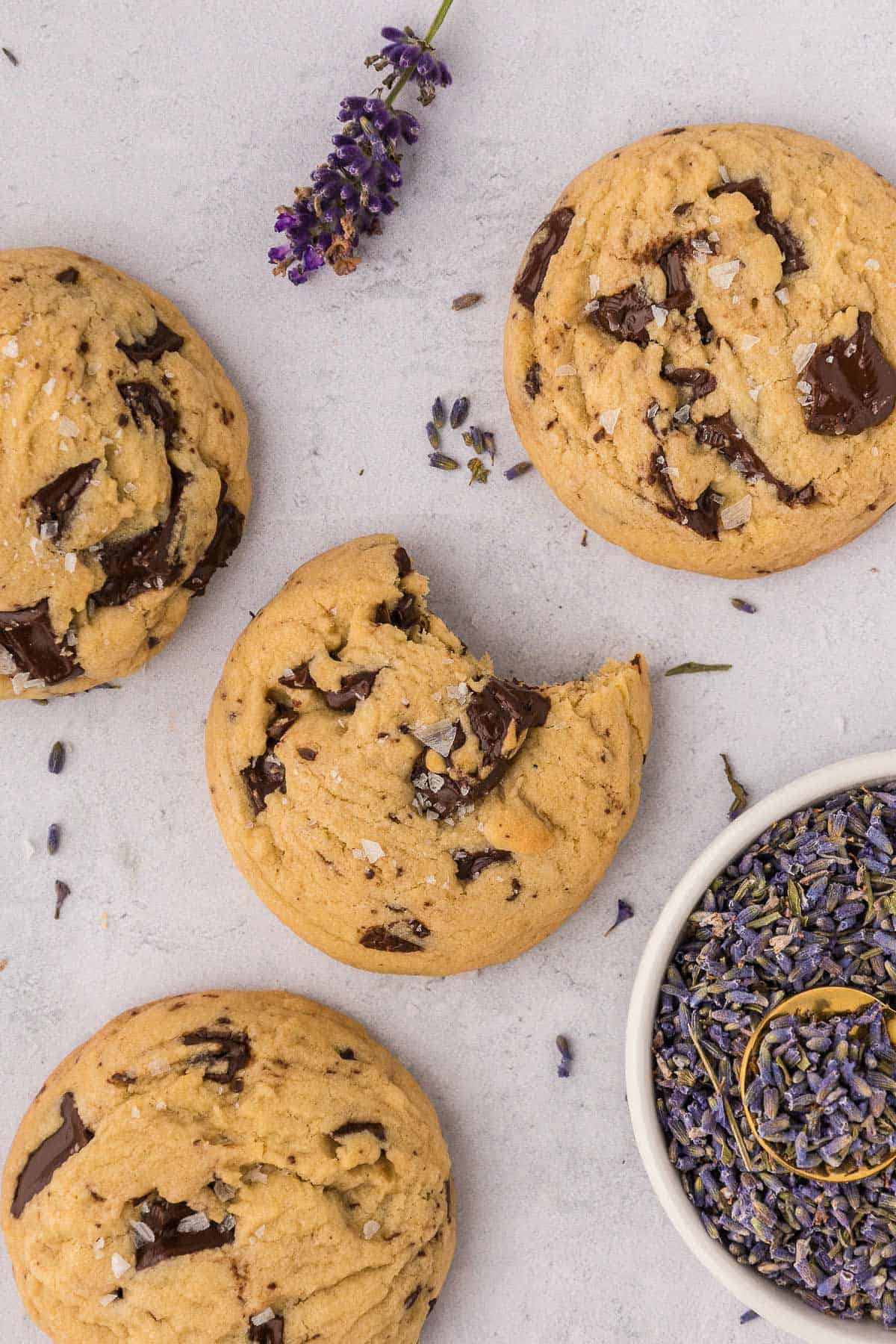 Four chocolate chunk cookies against a pink background with one bitten into and a bowl of lavender on the right.