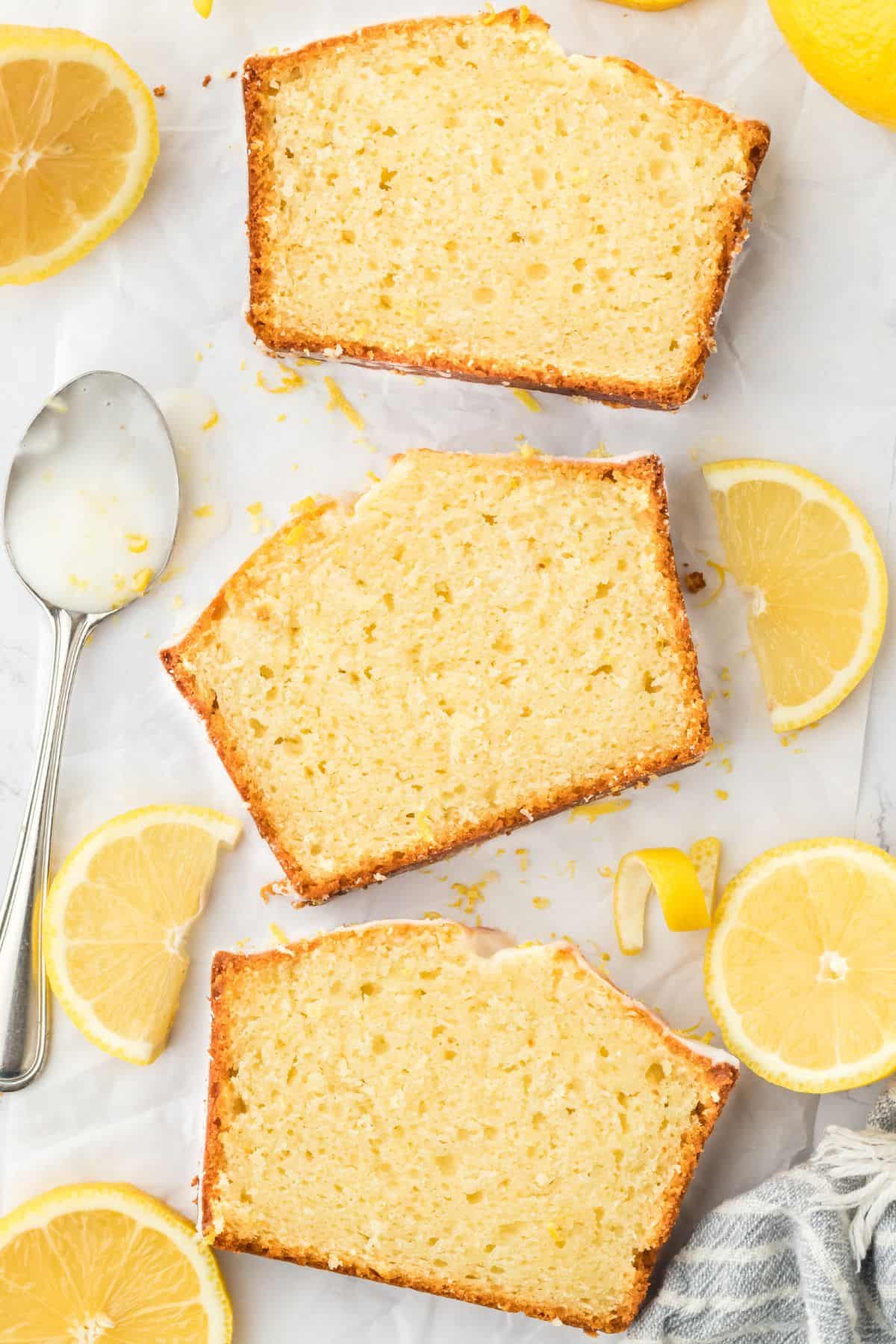Slices of lemon loaf cake laying on parchment on the table with lemons and slices around it.