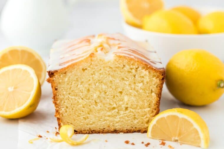 A lemon loaf cake sitting on the table surrounded by whole and halved lemons and a slice cut off to show the inside of the cake.