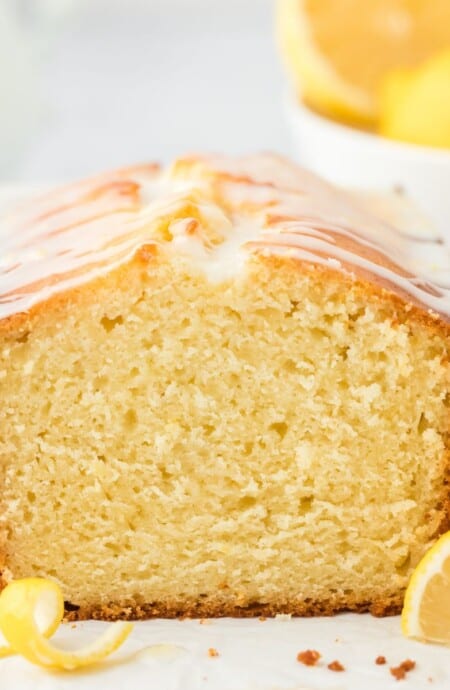 A lemon loaf cake on the table with the end piece cut to show the inside and a bowl of lemons in the background.