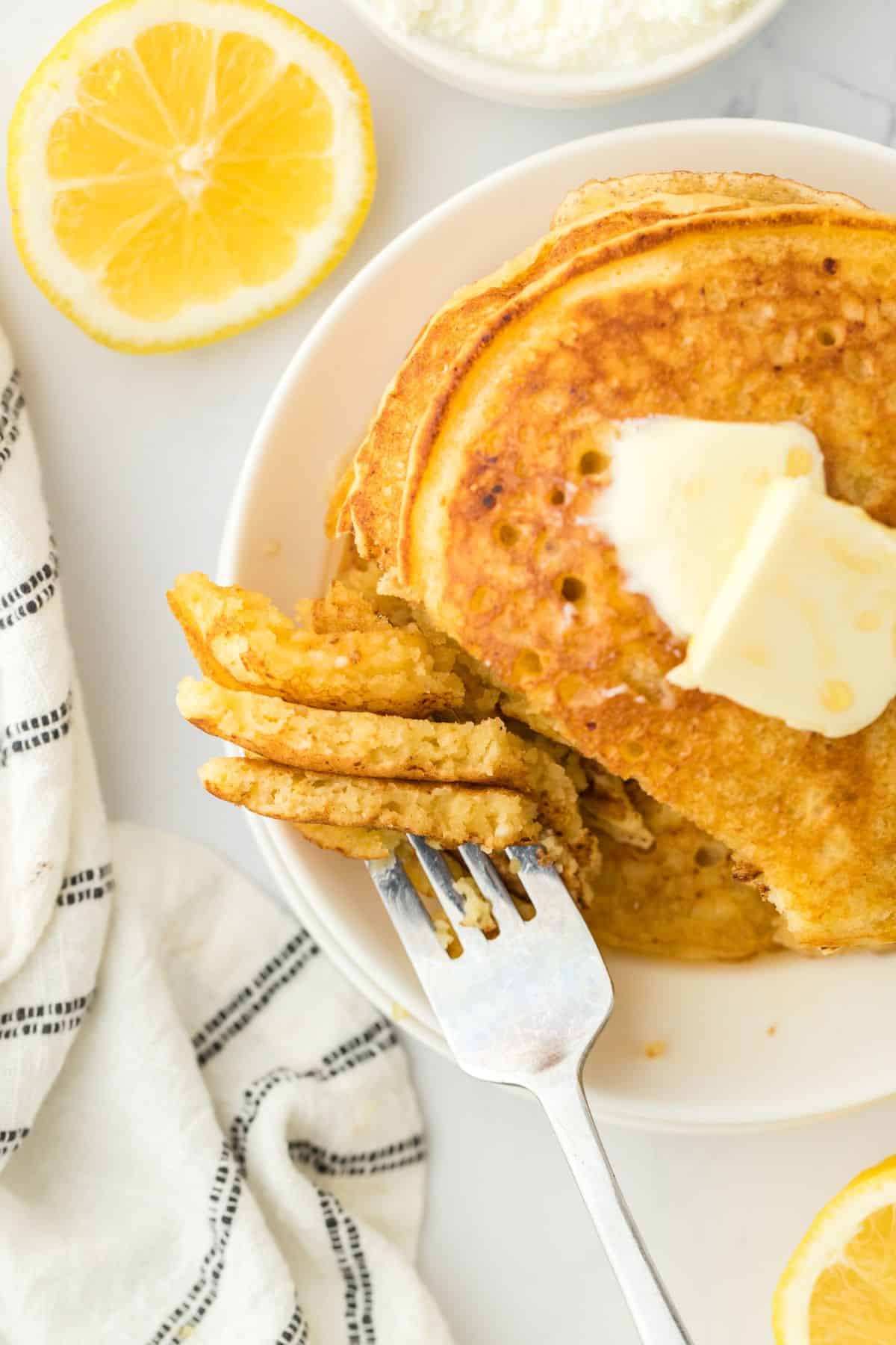 Ricotta Pancakes stacked high with a pat of butter on white plate with butter and lemons in background