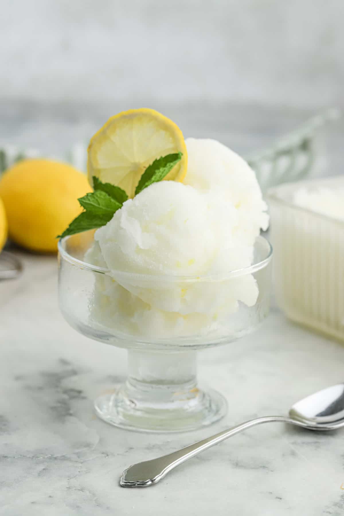 A glass dish of lemonade sorbet on the table with a piece of lemon slice and fresh mint for garnish.