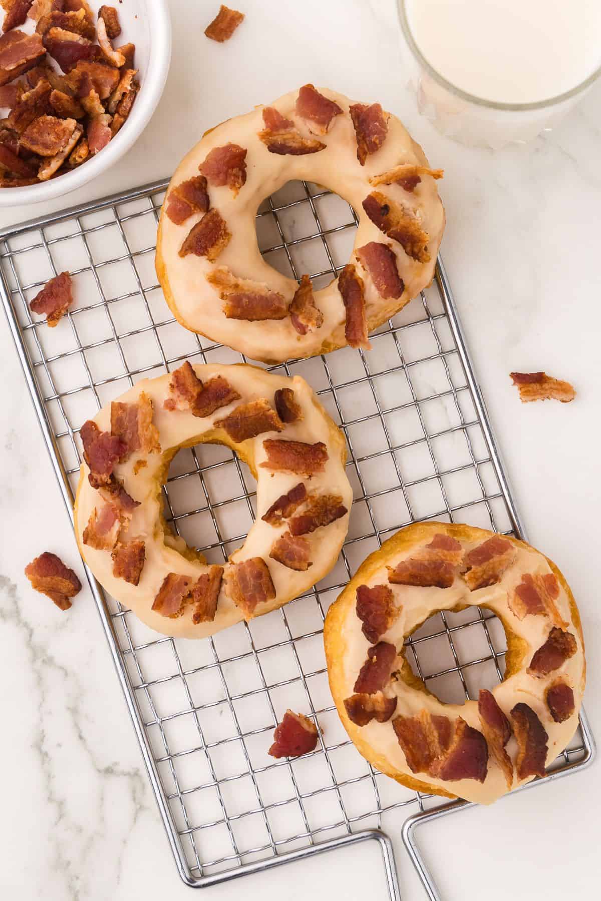 Maple bacon donuts on a wire rack with a glass of milk in the corner and a small bowl of bacon.