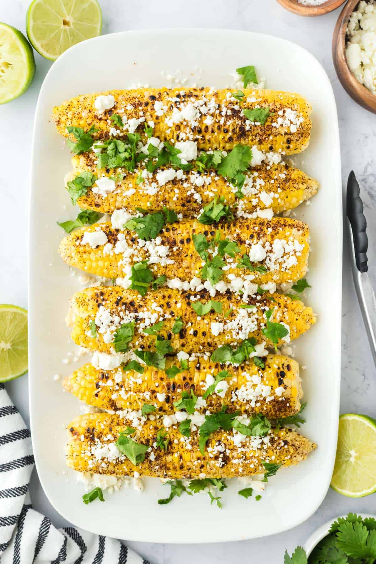 Overhead shot of Mexican corn on the cob on a white plate with limes and bowls of cheese around.