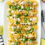 A platter of Mexican corn on the cob on the table topped with cilantro and white crumbled cheese.