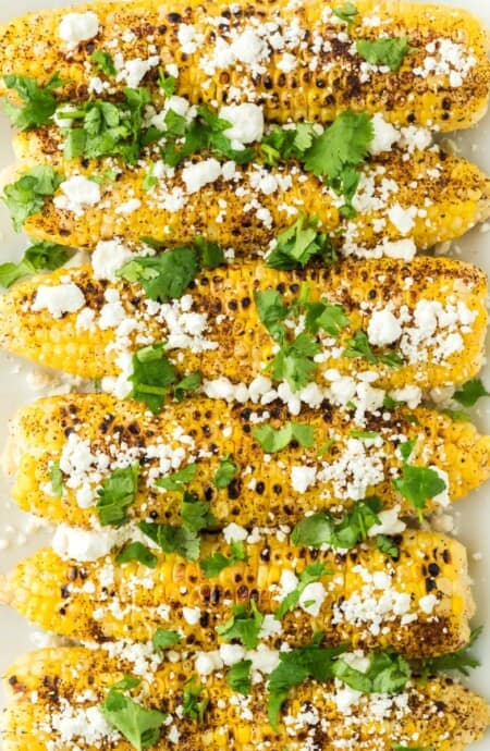 A platter of Mexican corn on the cob on the table topped with cilantro and white crumbled cheese.