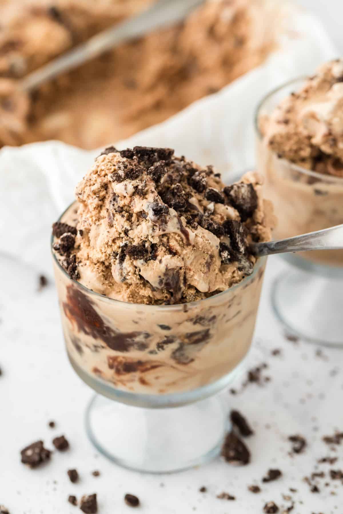 A glass dish of mocha fudge ice cream on a table with a spoon in the glass and cookies scattered on the table.