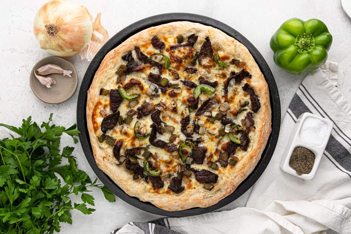 An overhead image of a steak pizza on the table with whole onion and bell pepper to the side.