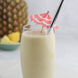 A glass of pina colada smoothie on the table with a straw and a drink umbrella in it.
