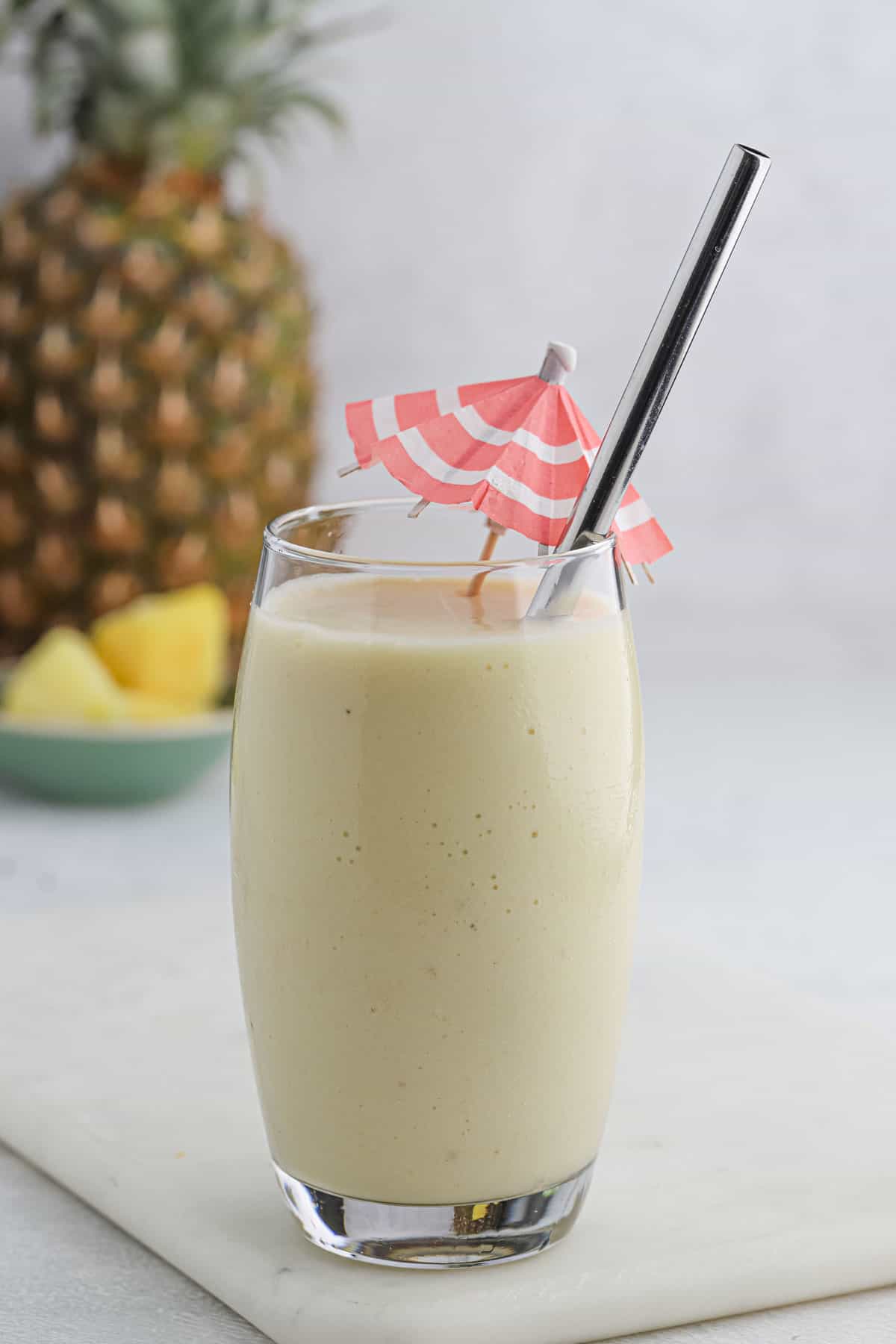 A glass of pina colada smoothie on the table with a straw and a drink umbrella in it.