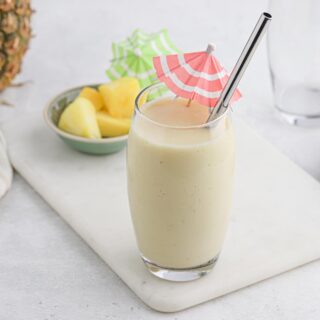 Pina colada smoothie in a glass on a white cutting board with a bowl of pineapple in the background.