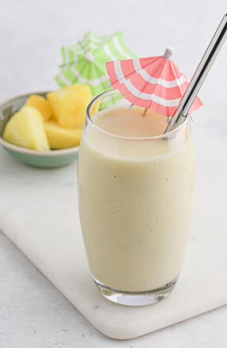 Pina colada smoothie in a glass on a white cutting board with a bowl of pineapple in the background.