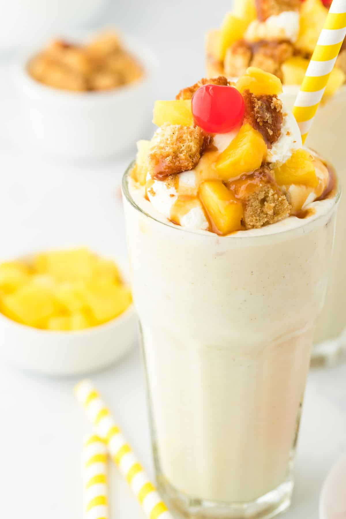 Two pineapple milkshakes with yellow and white straws in them and pineapple wedges and straws decorated around them.