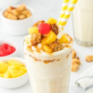 Pineapple milkshake in a glass with two straws and a bowl of toppings in the background.