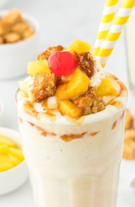 Pineapple milkshake in a glass with two straws and a bowl of toppings in the background.