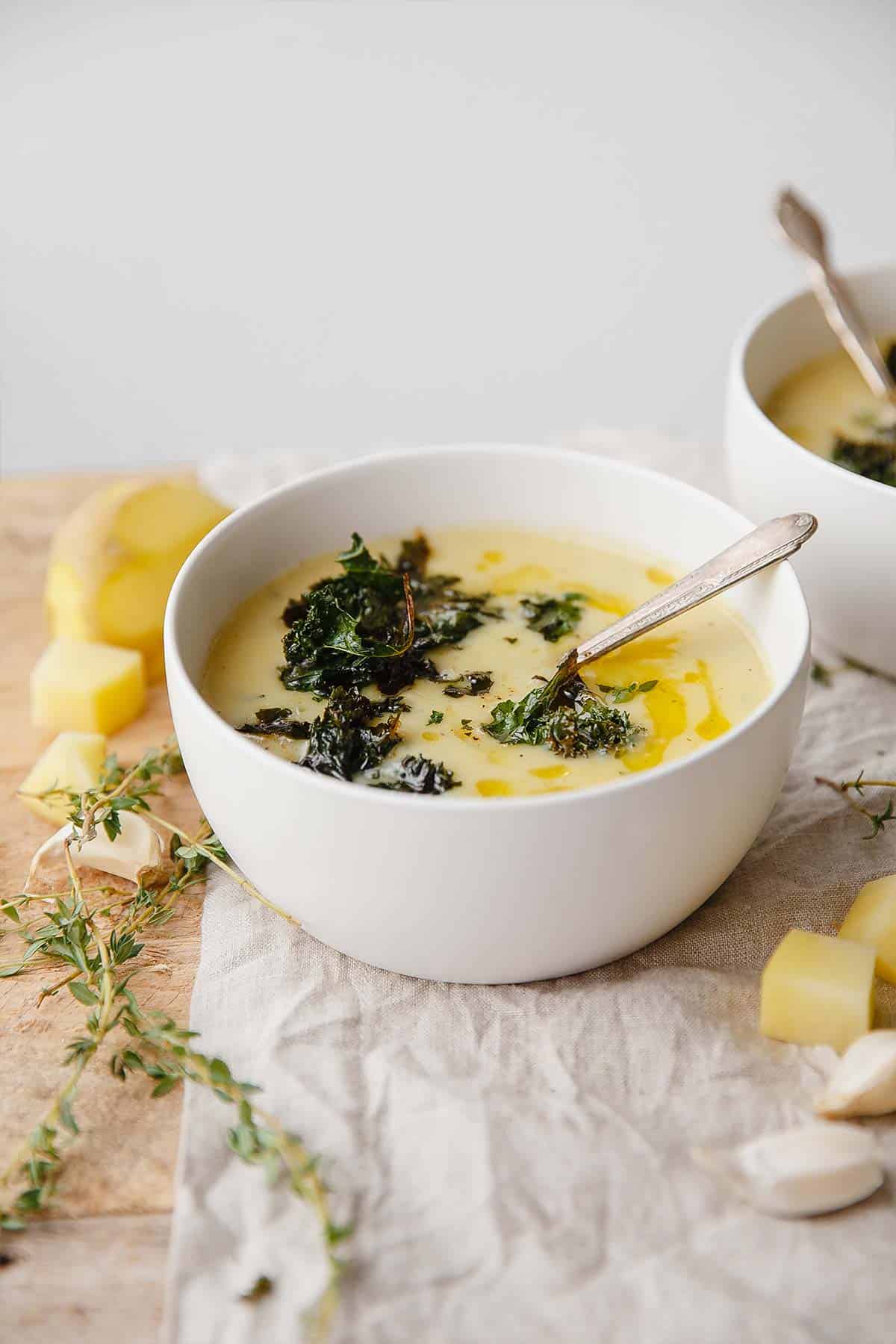 A bowl of potato and kale soup on the table with a spoon in it and thyme leaves, cheese, and garlic around the bowl on the table.