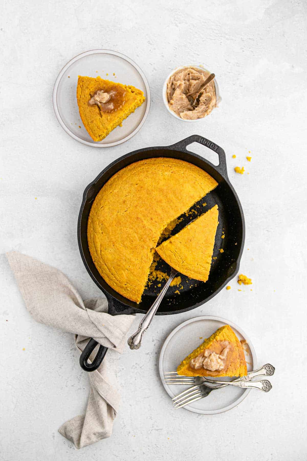 A cast iron skillet filled with pumpkin cornbread with a slice on a plate, one in the skillet and a bowl of cinnamon butter.