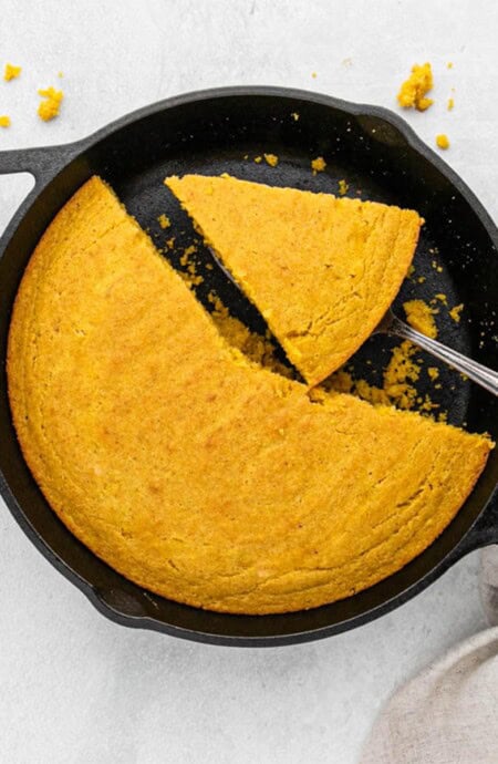 A cast iron skillet filled with pumpkin cornbread that is sliced and some pieces are missing.