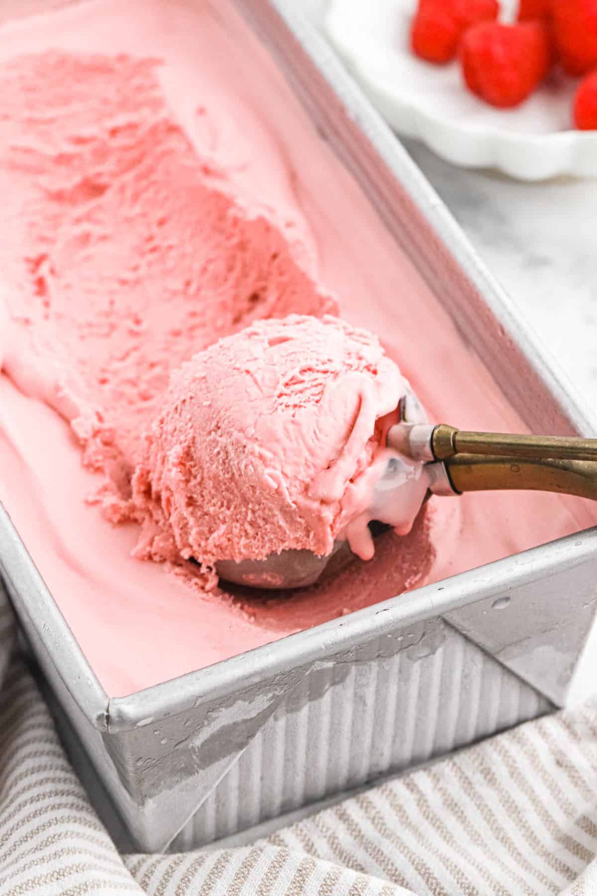 Large tray of raspberry ice cream with an ice cream scooper filled with a scoop of ice cream in it.