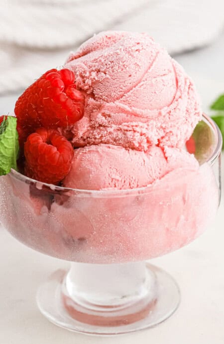 Raspberry ice cream scoops in a glass dessert bowl with fresh raspberries and mint.