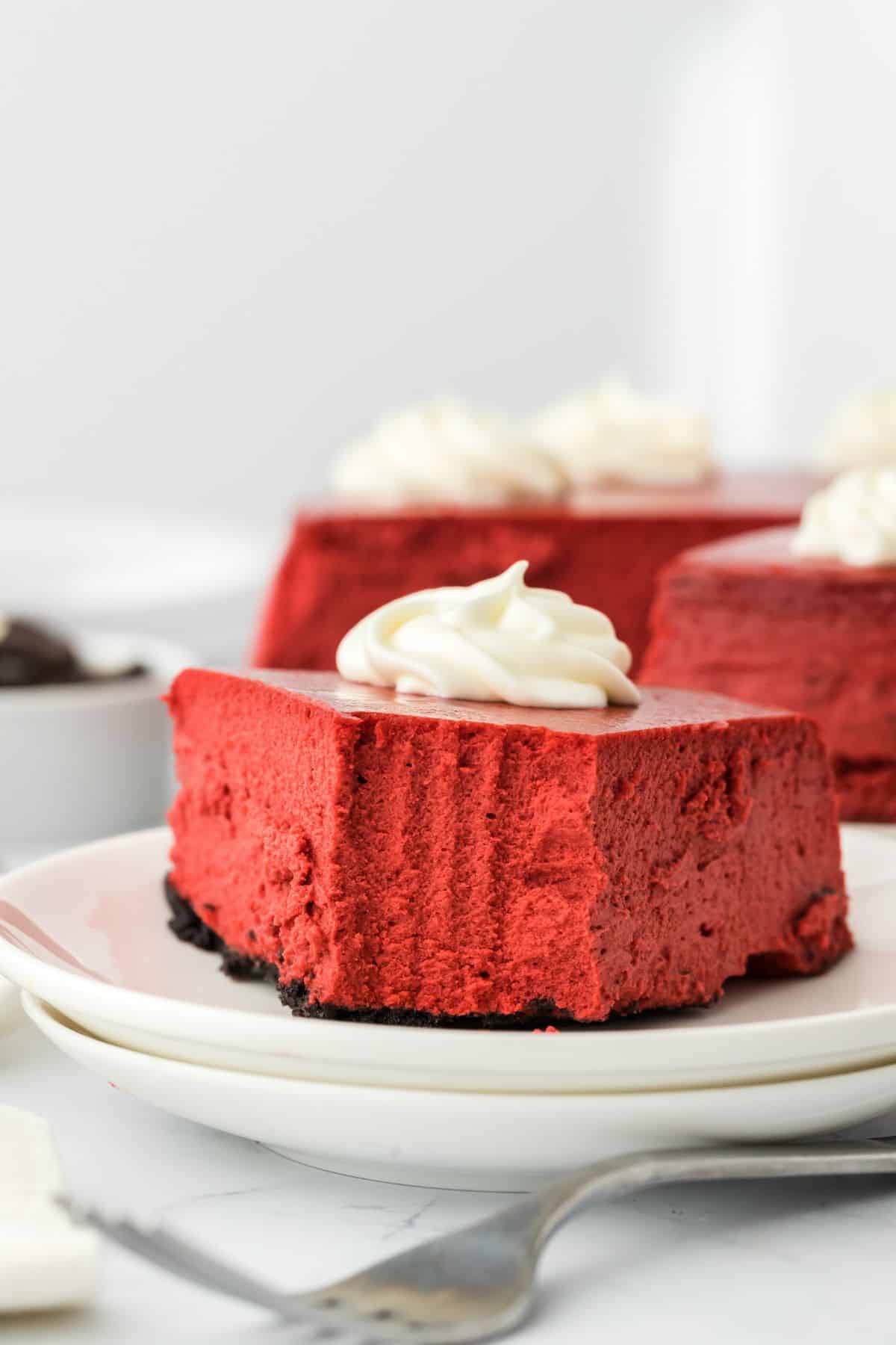 A slice of red velvet cheesecake on a stack of two plates topped with whipped cream and a bite missing.