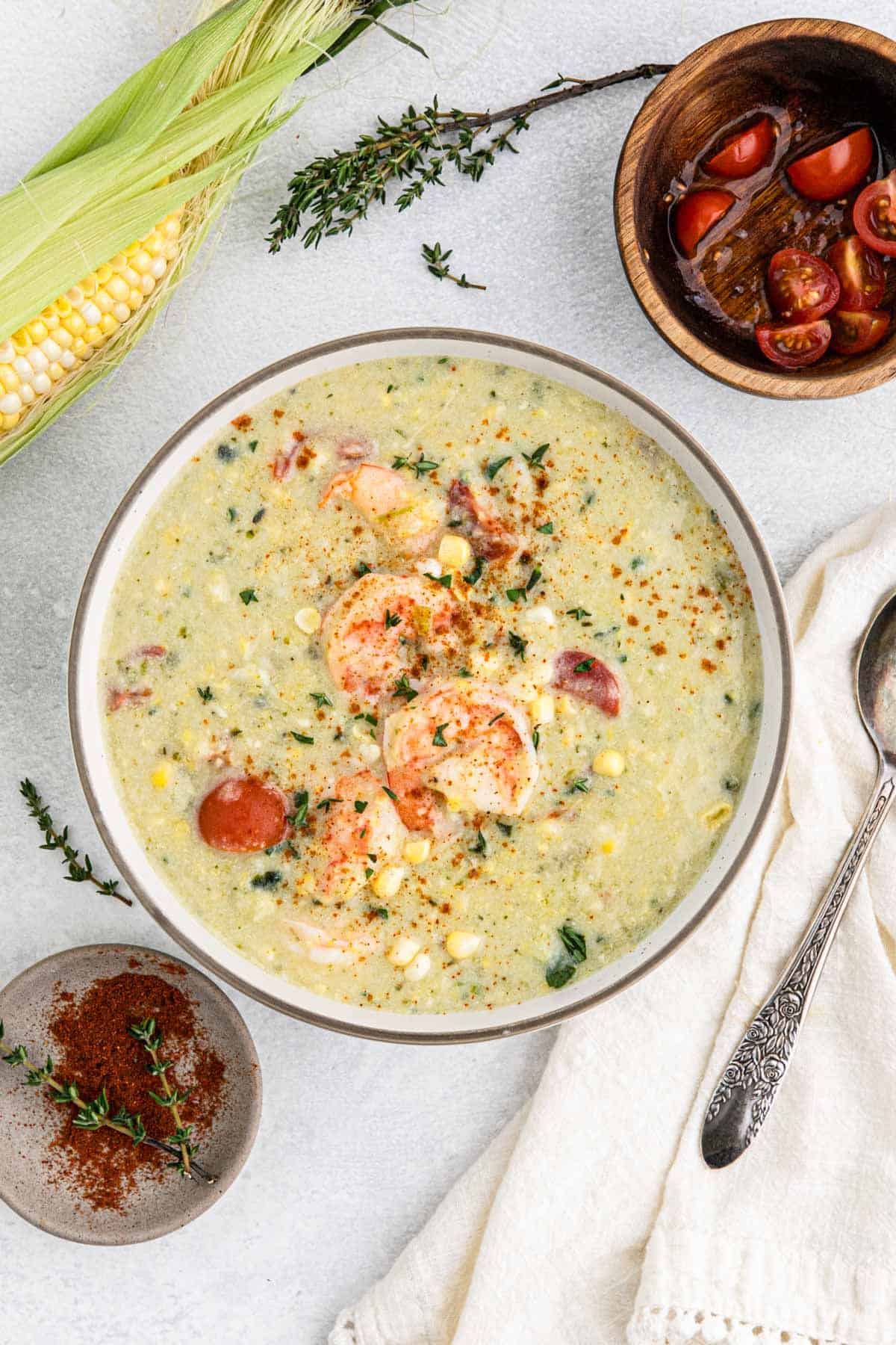 A large bowl of shrimp and corn chowder on the table with shrimp on top and a bowl of tomatoes and fresh corn on the side.