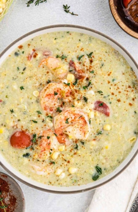 A bowl of shrimp corn chowder on the table with a spoon and napkin to the right and fresh ingredients around the bowl.