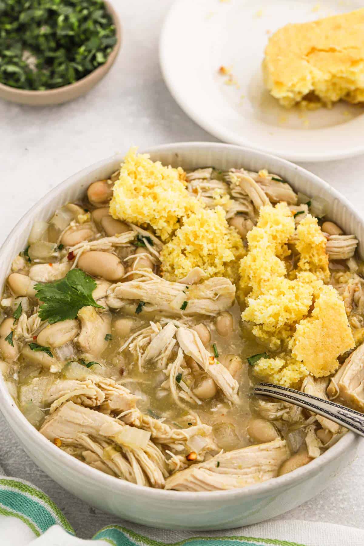 A bowl of crockpot chicken chili with crumbled cornbread on top is on the table with a bowl of cornbread and cilantro in the background.