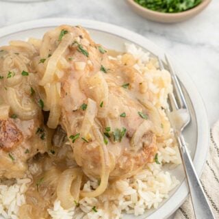 Southern smothered chicken with rice on a white plate with a fork to the side and a bowl of chopped parsley.
