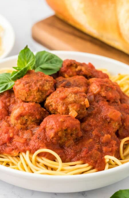 A plate of spaghetti and meatballs on the table garnished with a basil leaf and a loaf of bread behind.