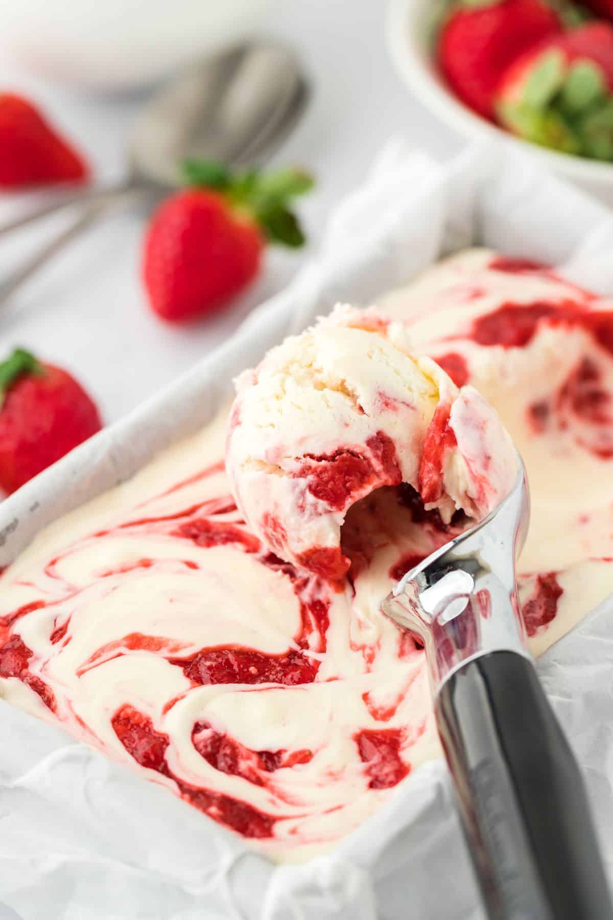 Scooping up strawberry cheesecake ice cream from a loaf pan after it's frozen that's surrounded by a bowl of strawberries and some on the table. 