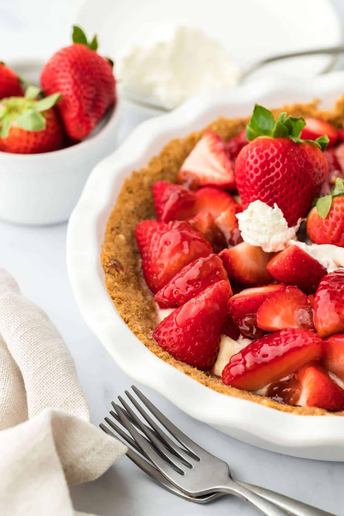 A strawberry tart on the table with only half of the dish showing and a bowl of strawberries in the background.
