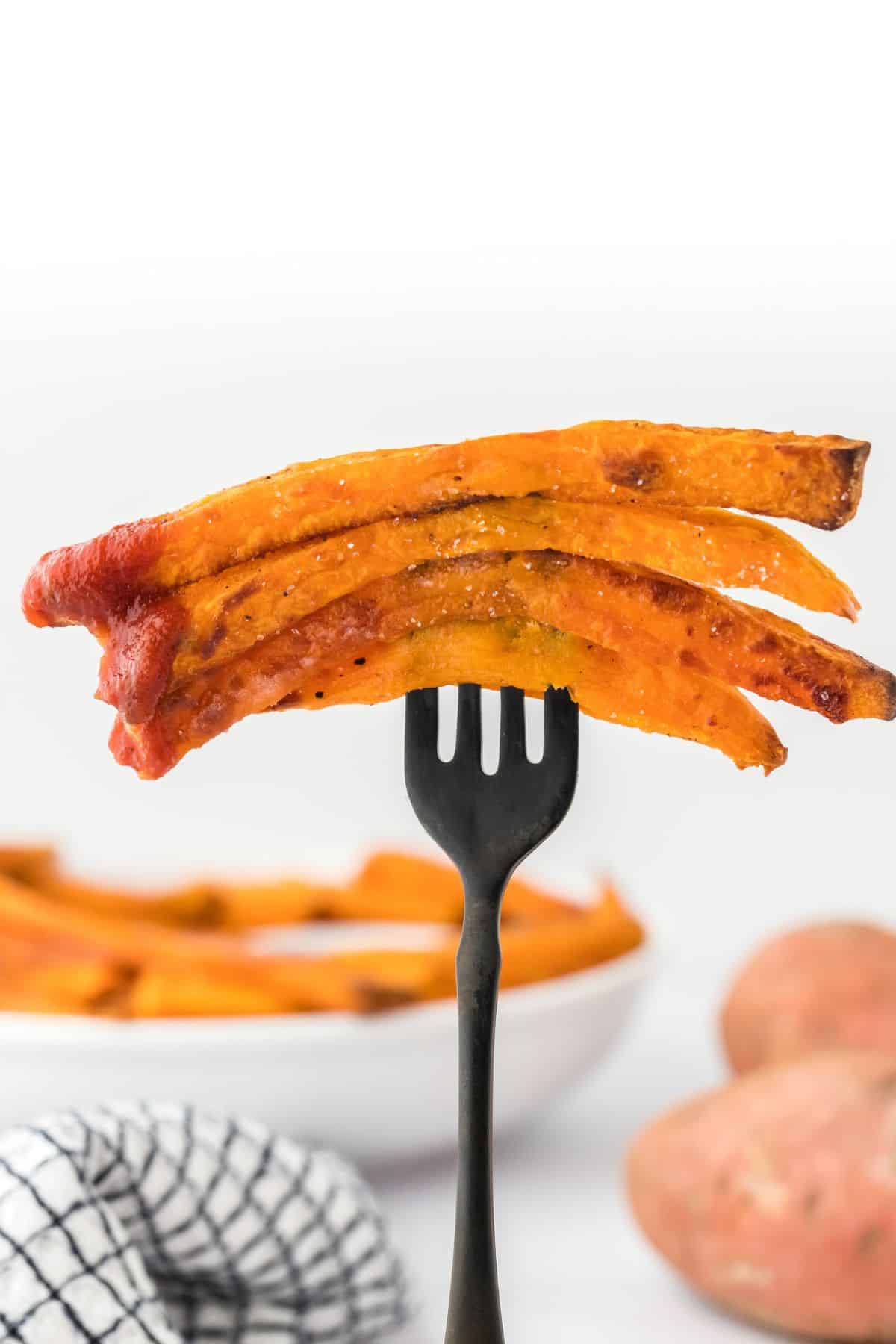 A fork held up in front of the table with four sweet potato fries on a fork in front of a bowl of more potatoes in the back.