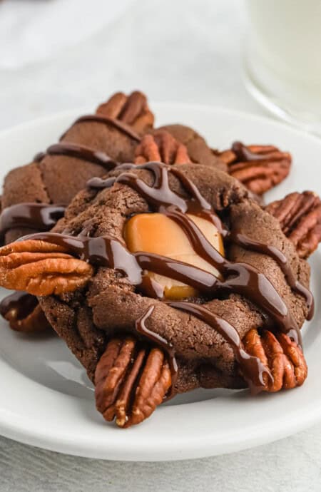 Two chocolate turtle cookies drizzled with chocolate sauce on a plate.