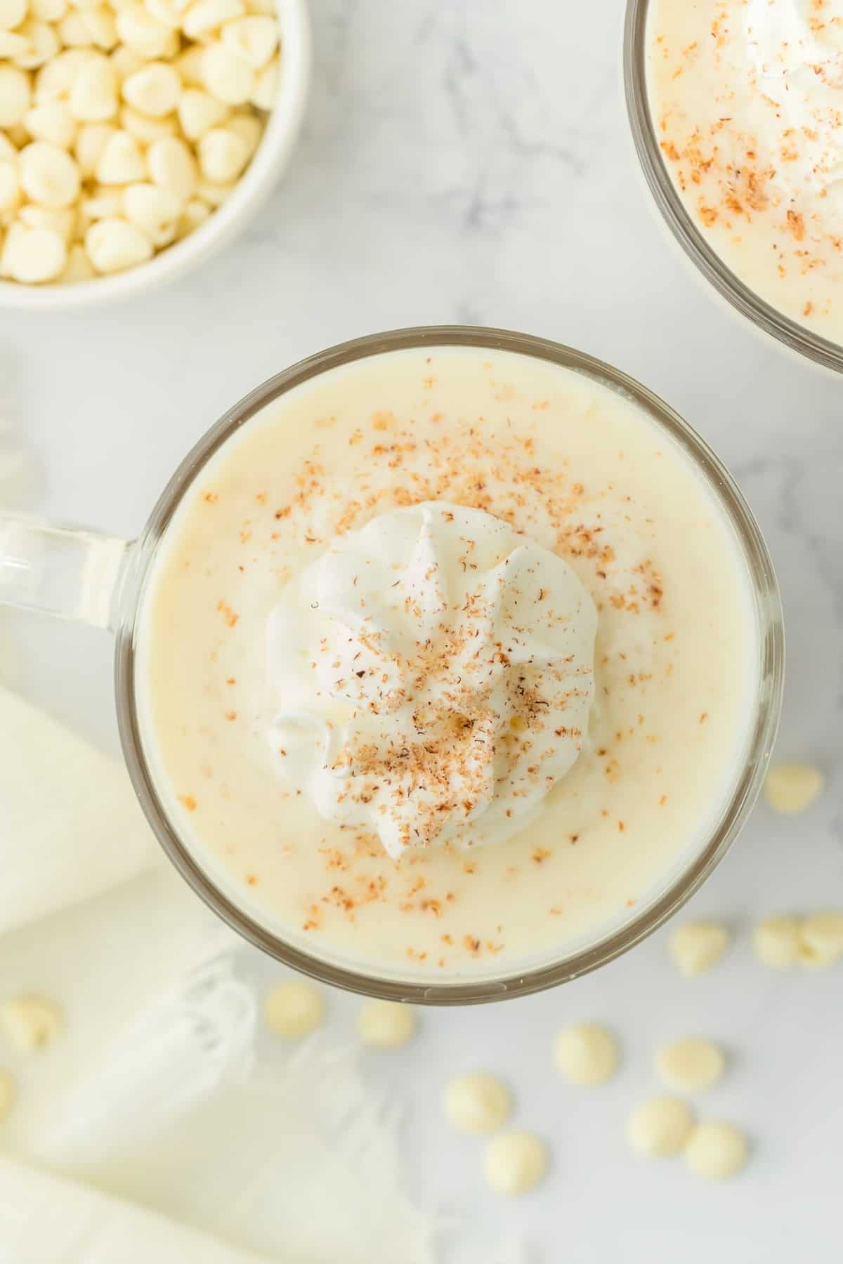 A clear mug filled with white chocolate hot chocolate topped with whipped cream and nutmeg.