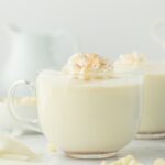 Mugs of white hot chocolate on the table in clear mugs with whipped cream on top.