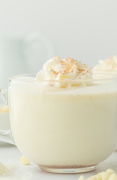 Mugs of white hot chocolate on the table in clear mugs with whipped cream on top.