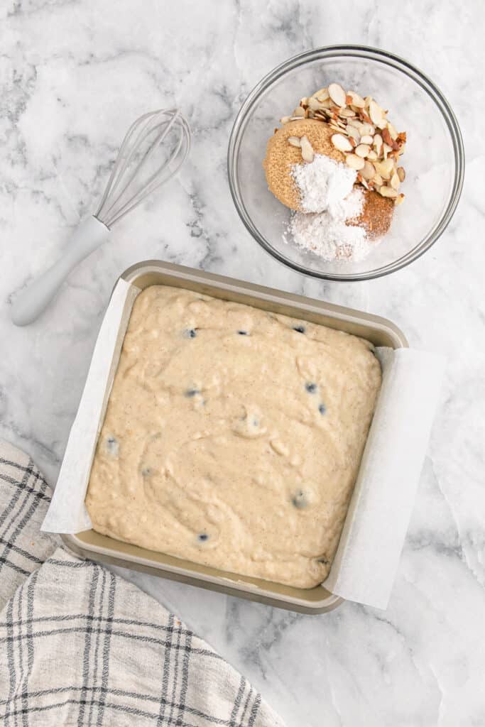 Blueberry cake batter in a square pan with crumble topping in a bowl