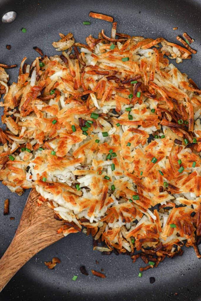 Cooked hash browns in a skillet with a wooden spoon under the potatoes in the pan.