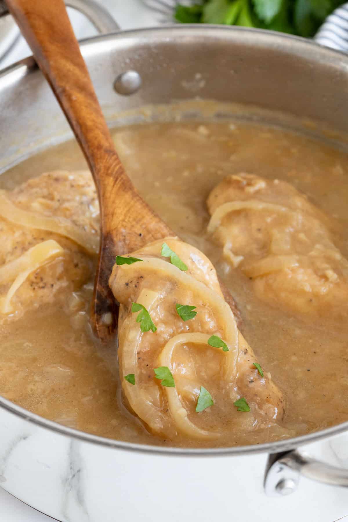 Closeup of a wooden spoon lifting a piece of Southern smothered chicken out of a pot of brown gravy, garnished with parsley