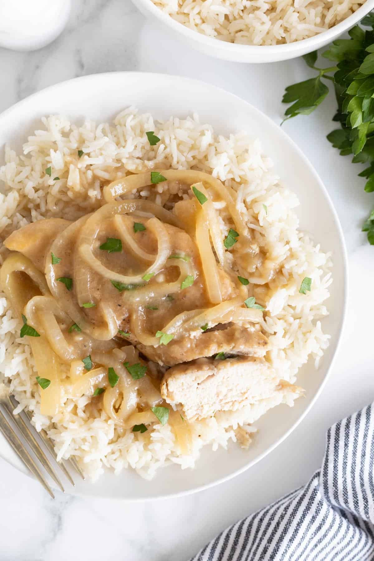 Overhead shot of a white plate with a serving of Southern smothered chicken over rice, with a piece of chicken cut open to show the white meat, and a fork beside it