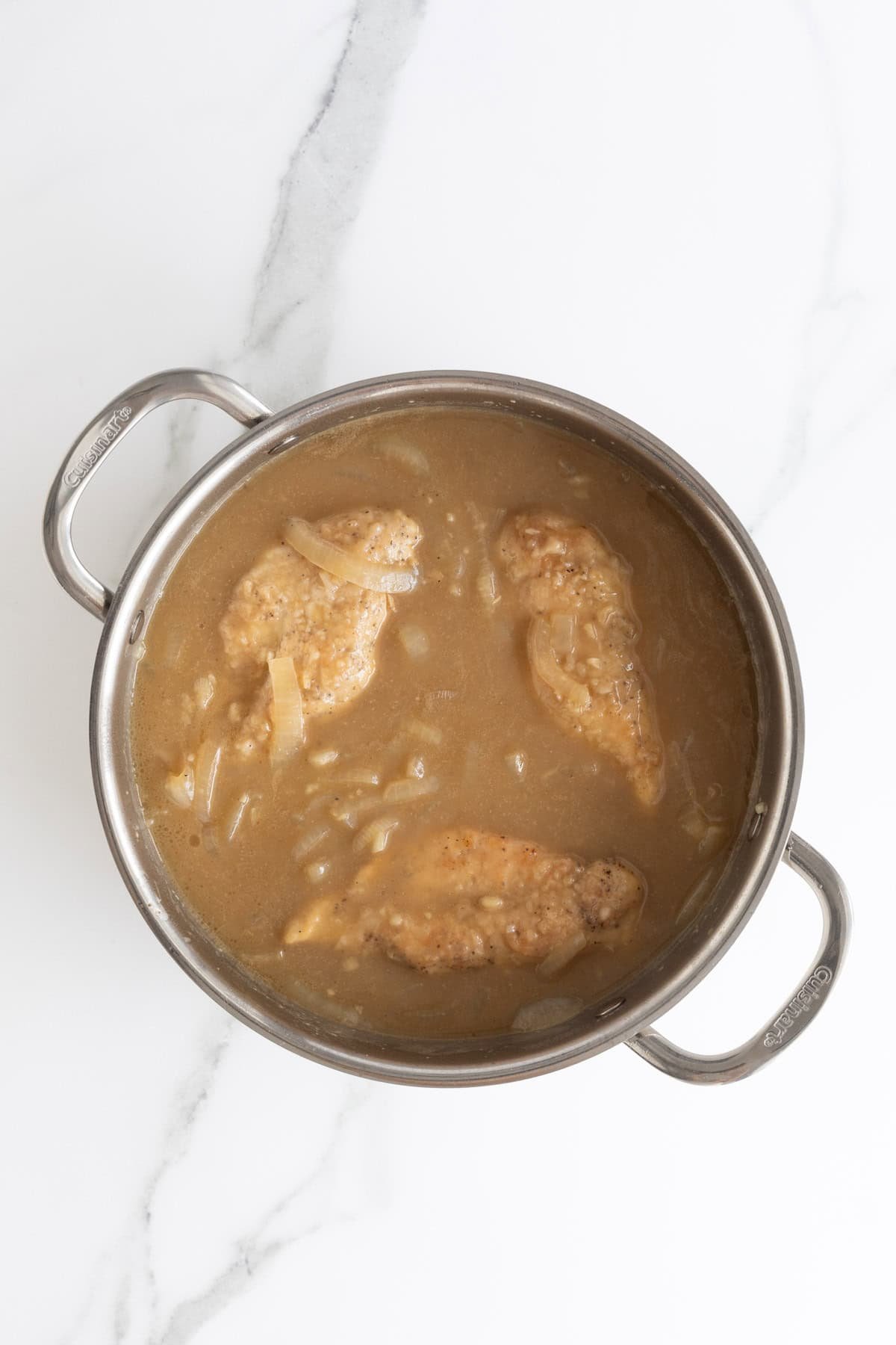 Three chicken breasts submerged in a thick, brown gravy with onions in a stainless steel pot