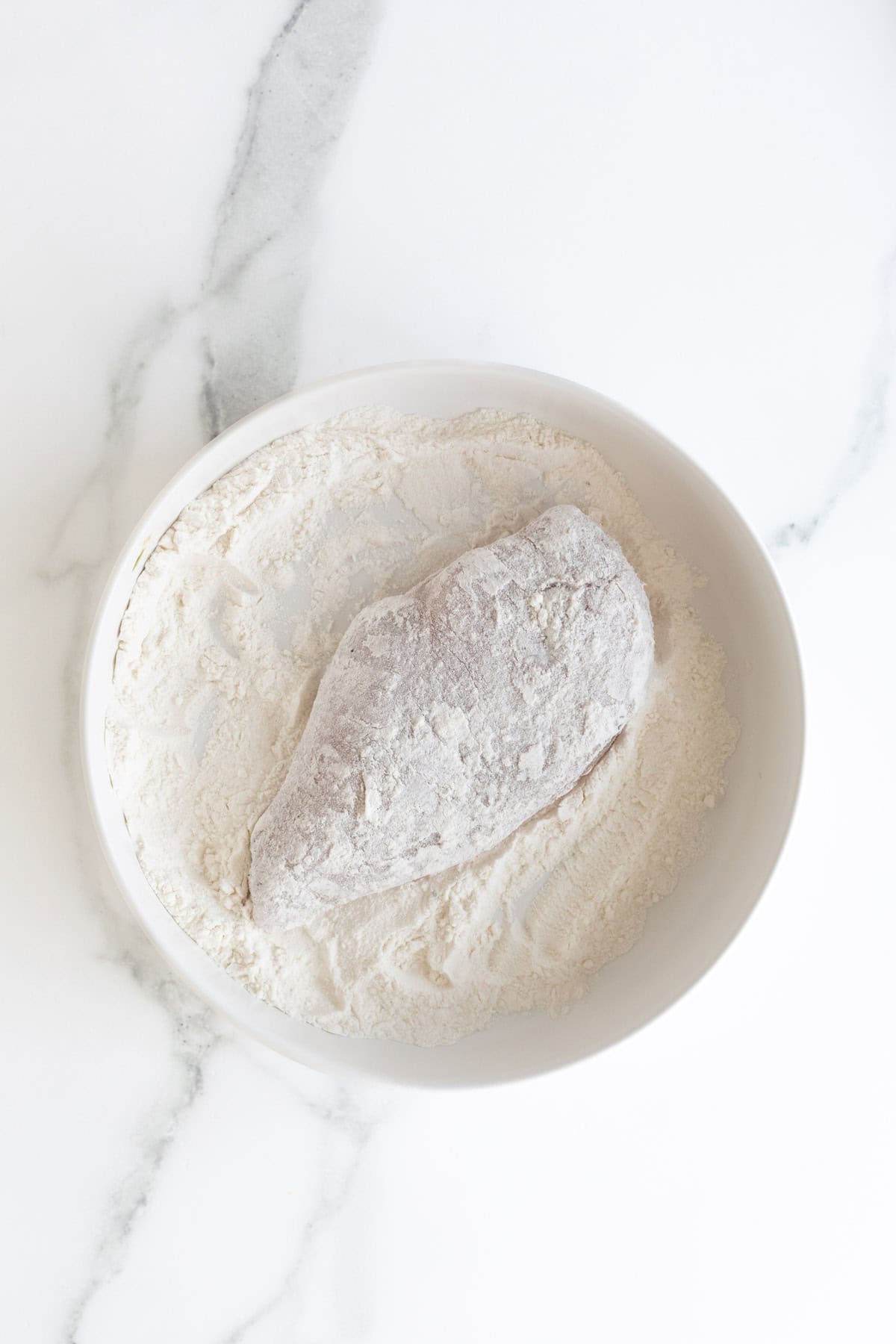 A chicken breast fully coated in white flour in a white bowl