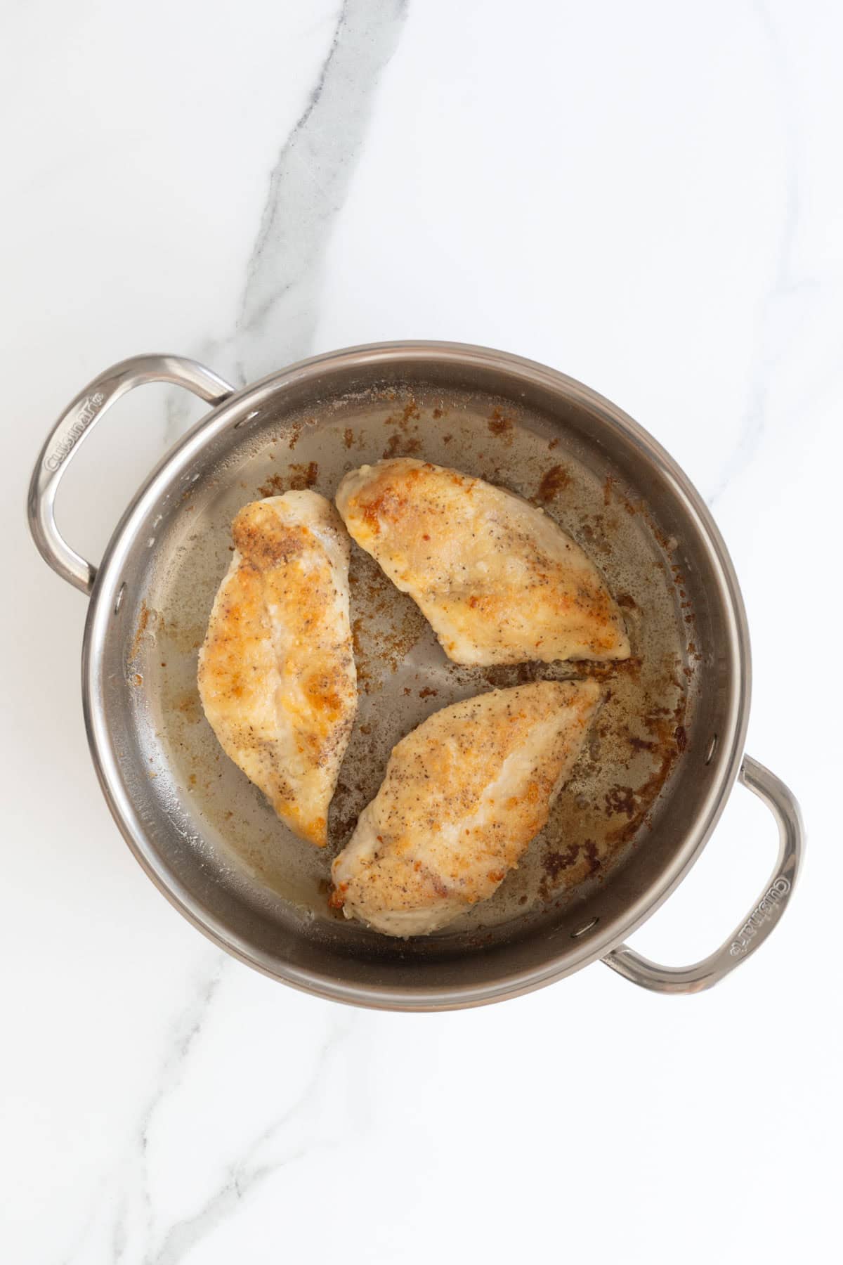 Three browned chicken breasts cooking in a stainless steel skillet